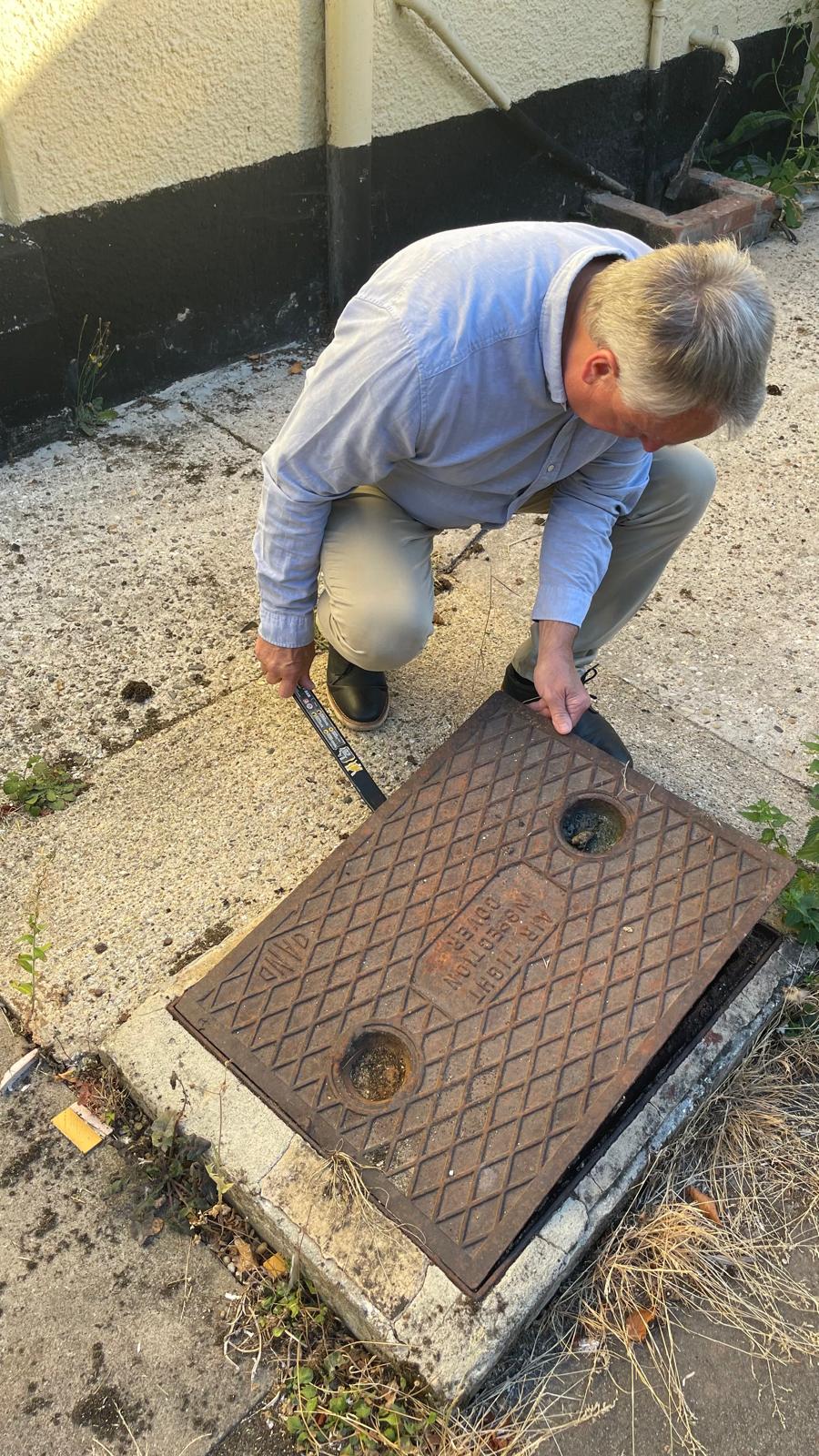 A surveyor kneels beside a weathered cast iron drainage cover during a property inspection in Norwich, Norfolk.